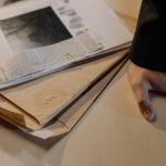 Mão sobre uma mesa ao lado de documentos de investigação e recortes de jornal.