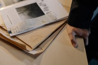 Mão sobre uma mesa ao lado de documentos de investigação e recortes de jornal.