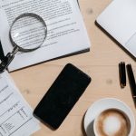 Documentos, laptop, café e lupa dispostos sobre uma mesa de escritório, vistos de cima.