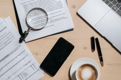 Documentos, laptop, café e lupa dispostos sobre uma mesa de escritório, vistos de cima.