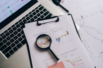 Vista superior de gráficos financeiros, um laptop e uma lupa sobre uma mesa, representando análise e investigação documental.