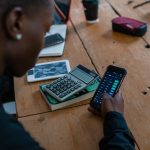 Pessoa analisa dados financeiros no celular, com uma calculadora sobre a mesa.